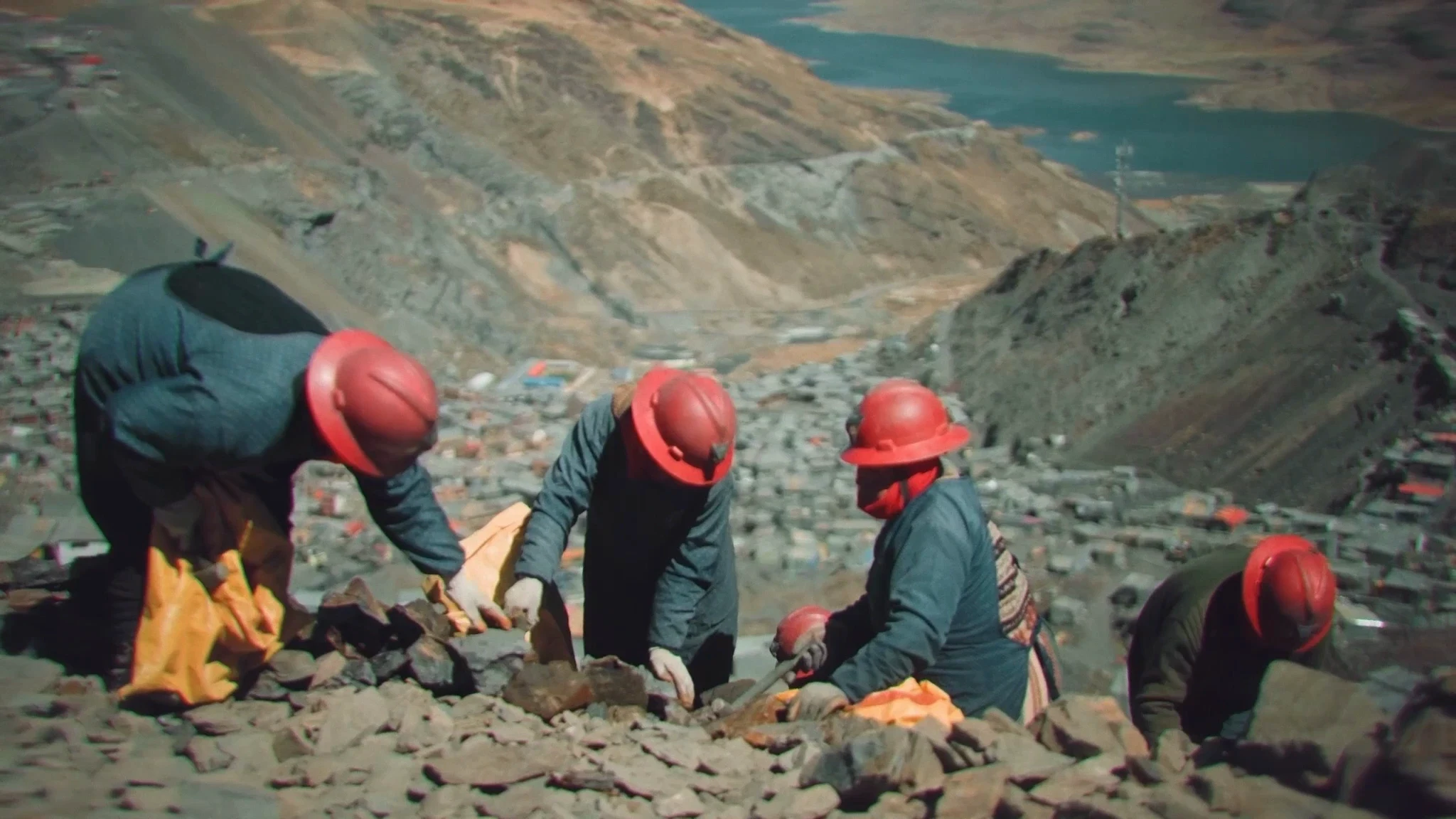 Mineras con casco rojo buscan mineral en la ladera, con una laguna andina al fondo